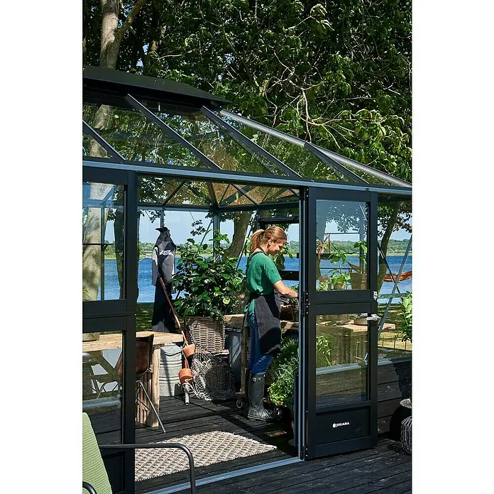 Juliana Gewächshaus Grand Oase - Außenmaß inkl. Dachüberstand (B x T): 296 x 439 cm, Material Seitenwand: Glas, 3 mm, Anthrazit/Schwarz - Frau im Juliana Gewächshaus mit schwarzem Rahmen. Ideal für Gartenliebhaber, bietet es Schutz und verlängert die Gartensaison.