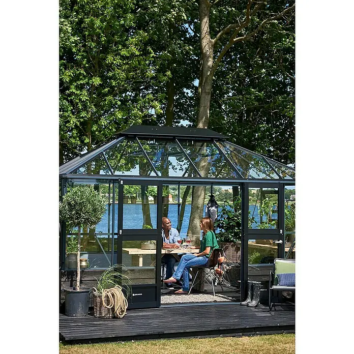 Juliana Gewächshaus Grand Oase - Außenmaß inkl. Dachüberstand (B x T): 296 x 439 cm, Material Seitenwand: Glas, 3 mm, Anthrazit/Schwarz - Ein Juliana Gewächshaus im Garten, in dem ein Paar am Tisch sitzt und Wein trinkt, umgeben von üppigem Grün.