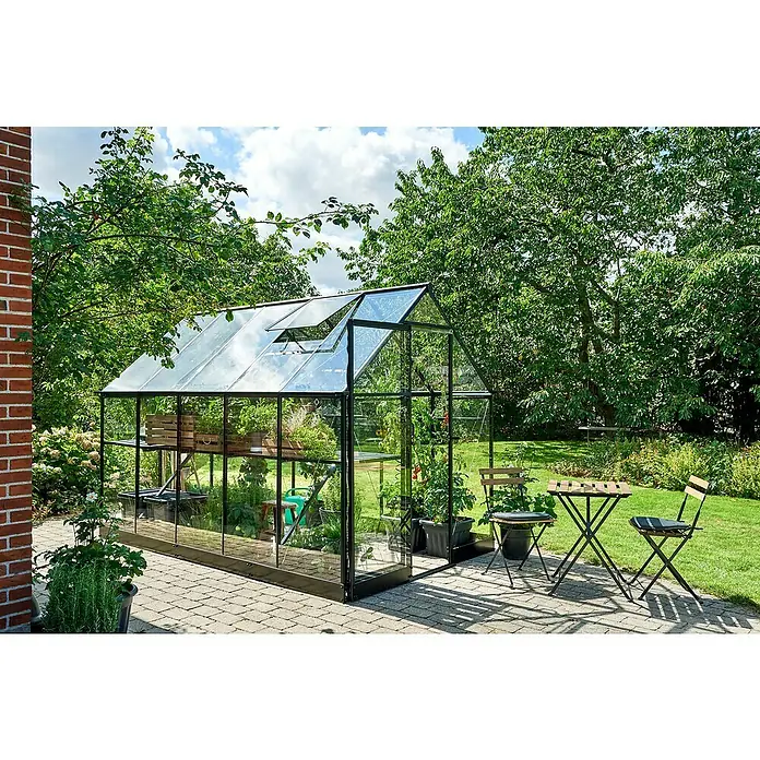 Halls Gewächshaus Qube 610 - Außenmaß inkl. Dachüberstand (B x T): 198 x 321 cm, Material Seitenwand: Glas, 3 mm, Schwarz - Das Bild zeigt ein Gewächshaus aus Glas und Metall mit Pflanzen darin und Gartenmöbeln davor, umgeben von Bäumen und Rasen.