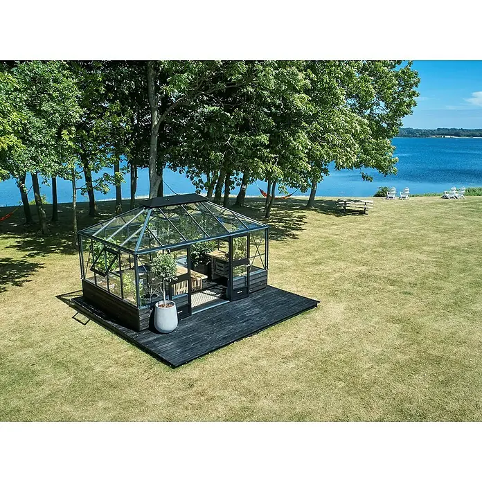 Juliana Gewächshaus Grand Oase - Außenmaß inkl. Dachüberstand (B x T): 296 x 439 cm, Material Seitenwand: Glas, 3 mm, Anthrazit/Schwarz - Das Gewächshaus mit seinem dunklen Holzdeck sticht auf dem Rasen hervor und bietet einen schönen Blick auf das blaue Wasser und die Bäume.