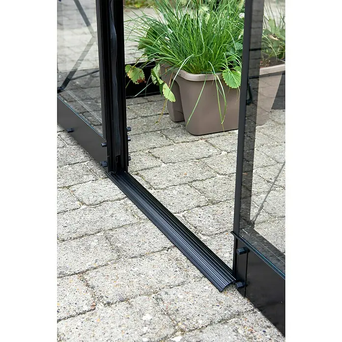 Halls Gewächshaus Qube 610 - Außenmaß inkl. Dachüberstand (B x T): 198 x 321 cm, Material Seitenwand: Glas, 3 mm, Schwarz - Nahaufnahme der schwarzen Aluminiumbasis eines Gewächshauses mit Glaswänden und Pflanzenkübeln auf einem gepflasterten Untergrund.