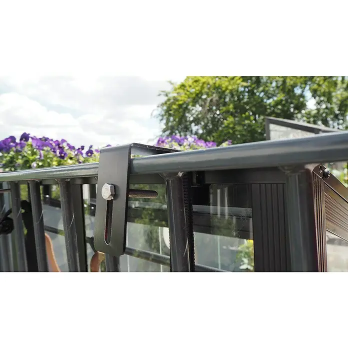 Juliana Gewächshaus Urban Balcony - Außenmaß inkl. Dachüberstand (B x T): 60 x 27 cm, Material Seitenwand: Glas, 4 mm, Schwarz - Nahaufnahme einer Blumenkastenhalterung aus Metall an einem Geländer mit Schrauben. Dahinter sind lila Blumen und grüne Blätter zu sehen.