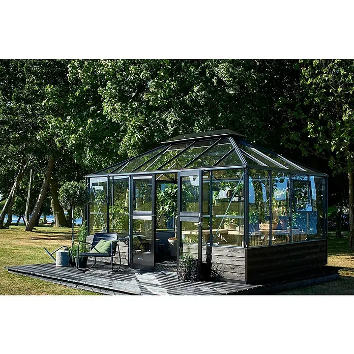 Juliana Gewächshaus Grand Oase - Außenmaß inkl. Dachüberstand (B x T): 296 x 439 cm, Material Seitenwand: Glas, 3 mm, Anthrazit/Schwarz - Das Bild zeigt ein Glashaus mit einer Holzterrasse und einem Stuhl davor. Das Glashaus steht im Grünen und ist von Bäumen umgeben.