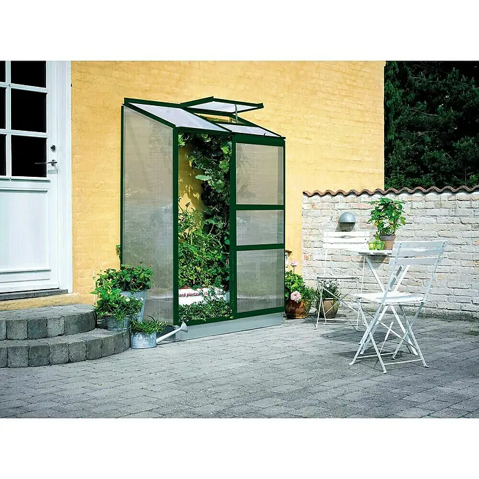 Halls Gewächshaus Altan 2 - Außenmaß inkl. Dachüberstand (B x T): 132 x 69 cm, Material Seitenwand: Kunststoff, 4 mm, Grün - Gewächshaus neben einer gelben Hauswand mit Pflanzen und einem Tisch mit Stühlen. Das grüne Gewächshaus bietet Platz für Gemüse und Kräuter.
