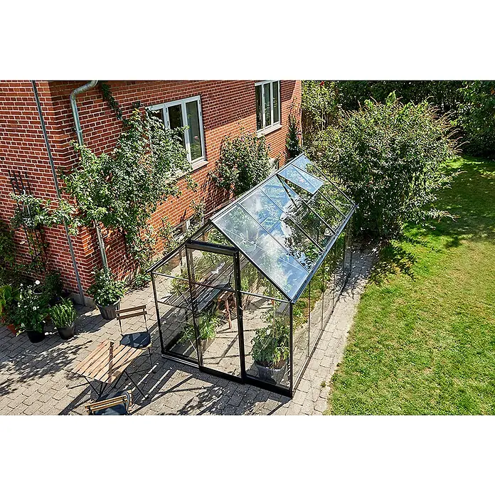Halls Gewächshaus Qube 610 - Außenmaß inkl. Dachüberstand (B x T): 198 x 321 cm, Material Seitenwand: Glas, 3 mm, Schwarz - Das Gewächshaus ist aus Glas mit einem schwarzen Rahmen, neben einem Haus aus rotem Backstein. Es hat zwei Dachfenster geöffnet.