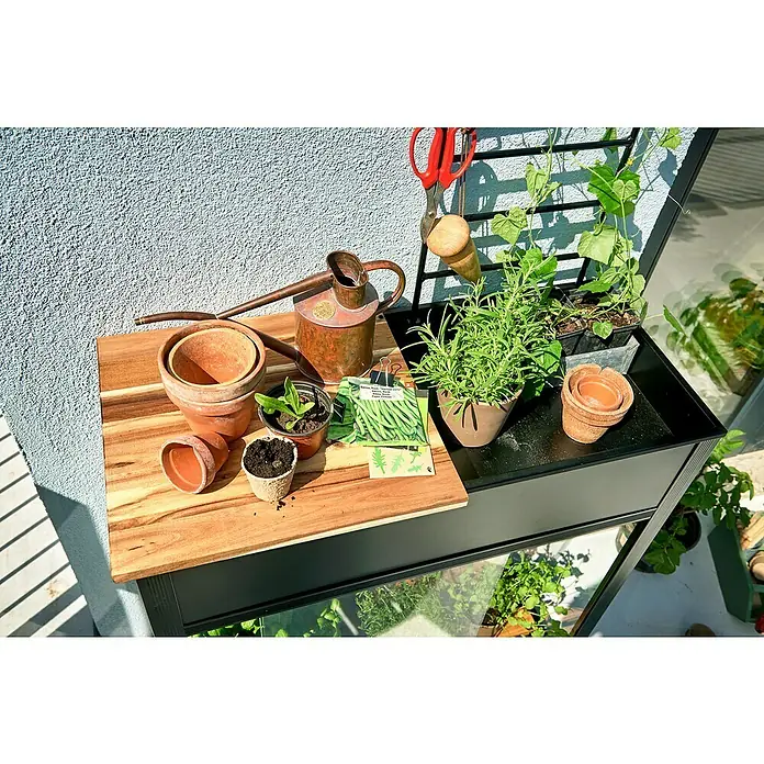 Juliana Gewächshaus Urban - Außenmaß inkl. Dachüberstand (B x T): 82 x 38 cm, Material Seitenwand: Glas, 4 mm, Schwarz - Nahaufnahme eines Gartentisches mit Pflanzen, Töpfen und Werkzeugen, ideal für Hobbygärtner und Balkonbepflanzung.