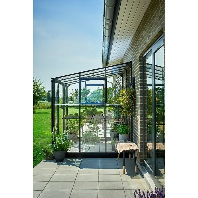 Halls Gewächshaus Qube Lean-to 68 - Außenmaß inkl. Dachüberstand (B x T): 251 x 188 cm, Material Seitenwand: Glas, Schwarz - Nahaufnahme eines Gewächshauses mit Pflanzen, das an ein Backsteingebäude angebaut ist, mit Sitzgelegenheiten im Freien auf einer Terrasse.