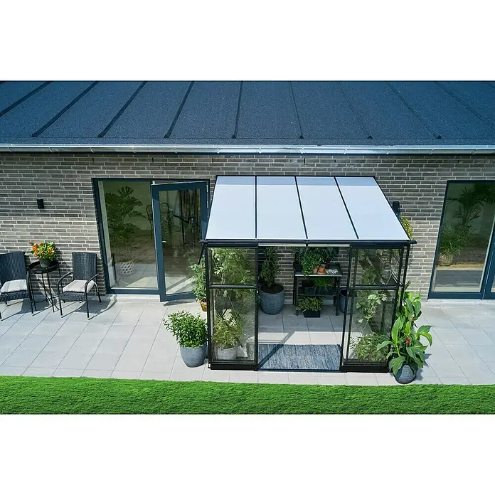Halls Gewächshaus Qube Lean-to 68 - Außenmaß inkl. Dachüberstand (B x T): 251 x 188 cm, Material Seitenwand: Glas, Schwarz - Das Bild zeigt ein Gewächshaus mit Pflanzen, das auf einer Terrasse neben einem Haus steht. Stühle und ein Tisch ergänzen die Szene.