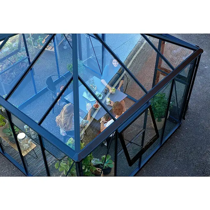 Juliana Gewächshaus Oase - Außenmaß inkl. Dachüberstand (B x T): 296 x 296 cm, Material Seitenwand: Glas, 3 mm, Anthrazit/Schwarz - Das Bild zeigt ein Gewächshaus mit Glaswänden, in dem sich Personen an einem Tisch aufhalten, umgeben von Pflanzen.