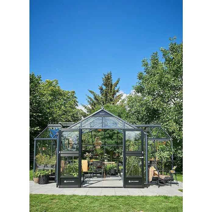 Juliana Gewächshaus Orangerie - Außenmaß inkl. Dachüberstand (B x T): 583 x 439 cm, Material Seitenwand: Glas, 3 mm, Anthrazit/Schwarz - Ein elegantes Juliana Gewächshaus mit offenem Eingang, umgeben von üppigem Grün und einem klaren, blauen Himmel.