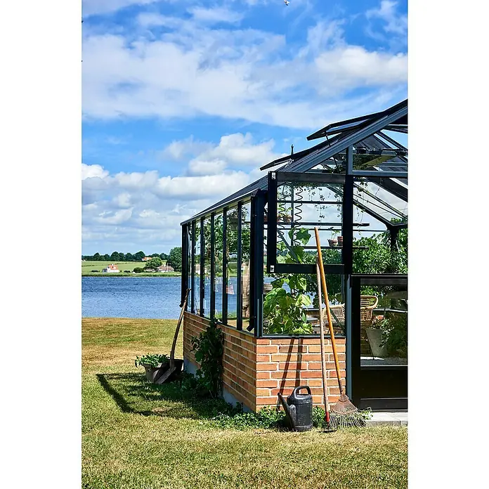 Gewächshaus Premium - Außenmaß inkl. Dachüberstand (B x T): 296 x 439 cm, Material Seitenwand: Glas, Anthrazit/Schwarz - Ein Gewächshaus mit Pflanzen und Gartengeräten im Garten, umgeben von einer idyllischen Landschaft, strahlt Ruhe und Naturnähe aus.