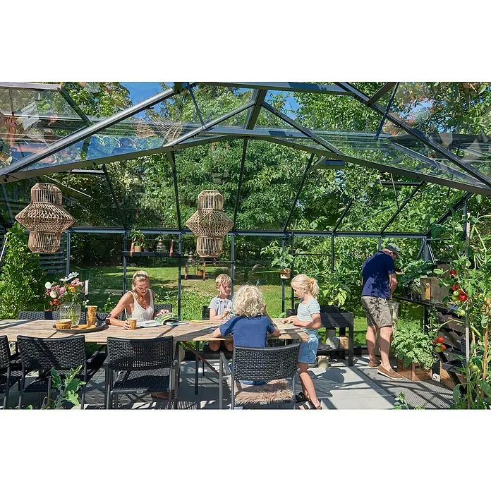 Juliana Gewächshaus Orangerie - Außenmaß inkl. Dachüberstand (B x T): 583 x 439 cm, Material Seitenwand: Glas, 3 mm, Anthrazit/Schwarz - Geräumiges Gewächshaus mit Familie am Tisch und einer Person, die sich um die Pflanzen kümmert, mit Hängeborden und einem grossen Tisch.