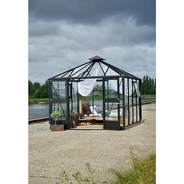 Juliana Gewächshaus Oase - Außenmaß inkl. Dachüberstand (B x T): 296 x 296 cm, Material Seitenwand: Glas, 3 mm, Anthrazit/Schwarz - Das Bild zeigt ein stilvolles Gartenhaus aus Glas mit einem Bett, das zum Entspannen einlädt. Ideal für ruhige Momente im Grünen.