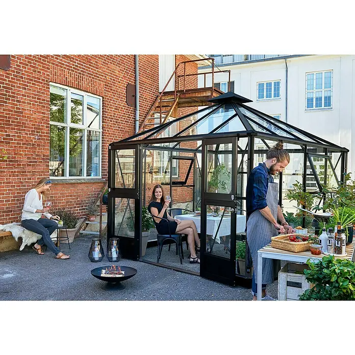 Juliana Gewächshaus Oase - Außenmaß inkl. Dachüberstand (B x T): 296 x 296 cm, Material Seitenwand: Glas, 3 mm, Anthrazit/Schwarz - Einladende Gartenszene mit einem Gewächshaus, in dem eine Frau sitzt, während draußen ein Mann kocht und eine weitere Frau entspannt.