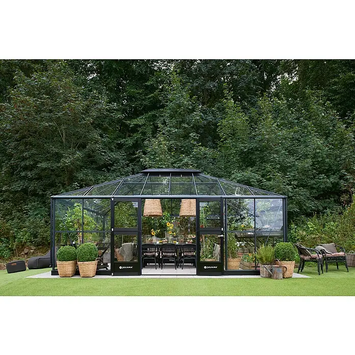 Juliana Gewächshaus Grand Oase - Außenmaß inkl. Dachüberstand (B x T): 439 x 583 cm, Material Seitenwand: Glas, 3 mm, Anthrazit/Schwarz - Ein Juliana Gewächshaus mit dunklem Rahmen im Garten. Es ist mit Tisch, Stühlen und Pflanzen geschmückt und bietet einen einladenden Raum.