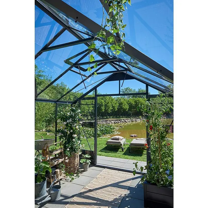 Juliana Gewächshaus Orangerie - Außenmaß inkl. Dachüberstand (B x T): 583 x 439 cm, Material Seitenwand: Glas, 3 mm, Anthrazit/Schwarz - Innenansicht eines Gewächshauses mit Pflanzen und Blick auf einen sonnigen Garten mit Liegestühlen und einem Teich.