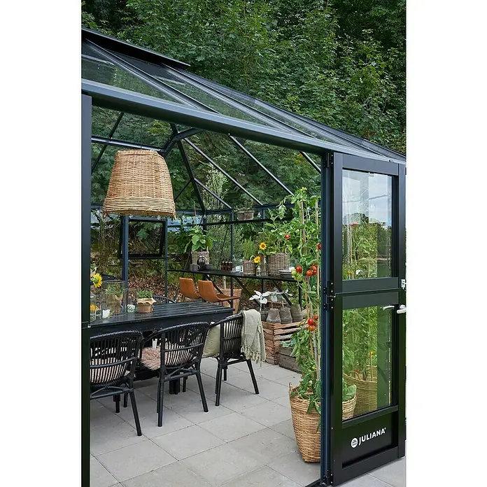 Juliana Gewächshaus Grand Oase - Außenmaß inkl. Dachüberstand (B x T): 439 x 583 cm, Material Seitenwand: Glas, 3 mm, Anthrazit/Schwarz - Ein Juliana Gewächshaus mit Esstisch und Stühlen, Pflanzen und einer hängenden Lampe. Ideal für Gartenliebhaber und stilvolle Entspannung.