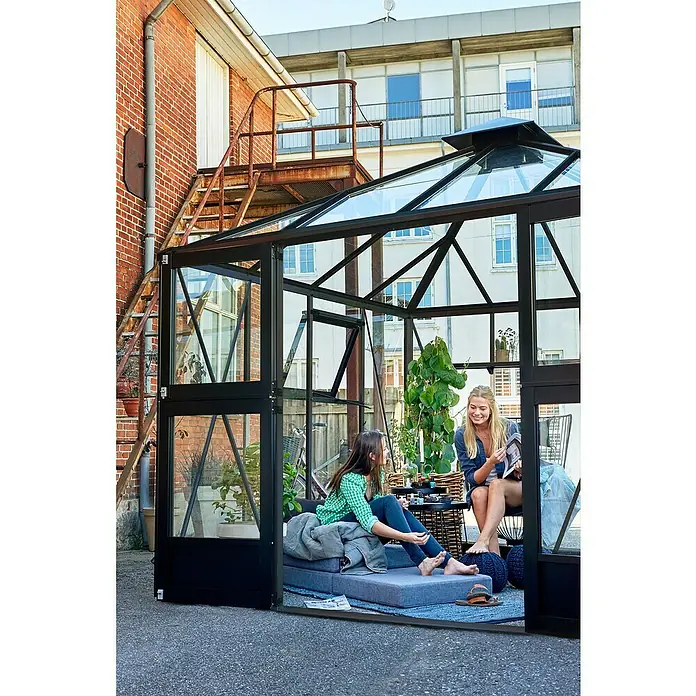 Juliana Gewächshaus Oase - Außenmaß inkl. Dachüberstand (B x T): 296 x 296 cm, Material Seitenwand: Glas, 3 mm, Anthrazit/Schwarz - Ein Gewächshaus mit zwei Frauen darin, die sich entspannen und lesen, eine grüne Pflanze im Hintergrund.