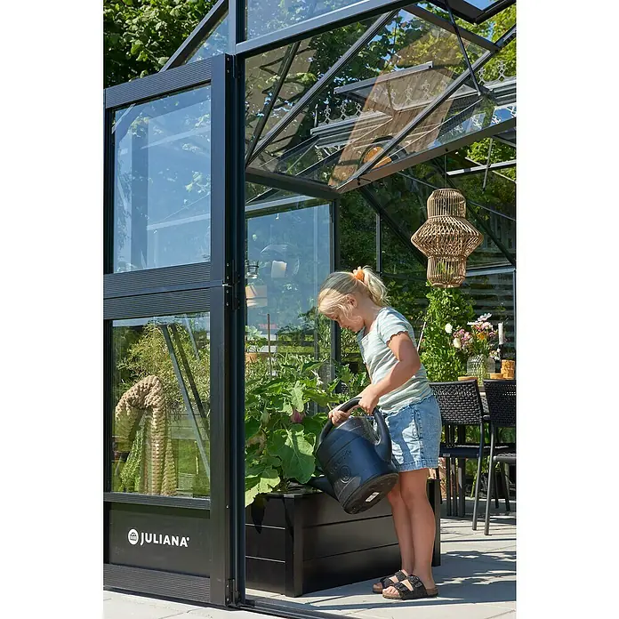 Juliana Gewächshaus Orangerie - Außenmaß inkl. Dachüberstand (B x T): 583 x 439 cm, Material Seitenwand: Glas, 3 mm, Anthrazit/Schwarz - Ein Mädchen gießt Pflanzen in einem Juliana Gewächshaus. Das Gewächshaus bietet optimalen Schutz für die Pflanzen.