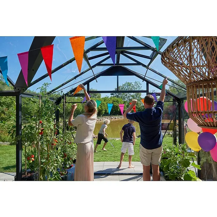 Juliana Gewächshaus Orangerie - Außenmaß inkl. Dachüberstand (B x T): 583 x 439 cm, Material Seitenwand: Glas, 3 mm, Anthrazit/Schwarz - Familie feiert im Gewächshaus, geschmückt mit bunten Wimpeln und Ballons, umgeben von Tomatenpflanzen und spielenden Kindern.