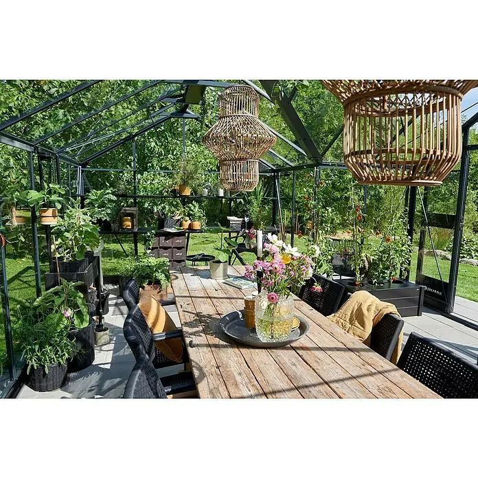 Juliana Gewächshaus Orangerie - Außenmaß inkl. Dachüberstand (B x T): 583 x 439 cm, Material Seitenwand: Glas, 3 mm, Anthrazit/Schwarz - Innenansicht eines Gewächshauses mit Holztisch, Stühlen und Pflanzen. Dekorative Lampen hängen von der Decke, während ein Blumenstrauß den Tisch schmückt.