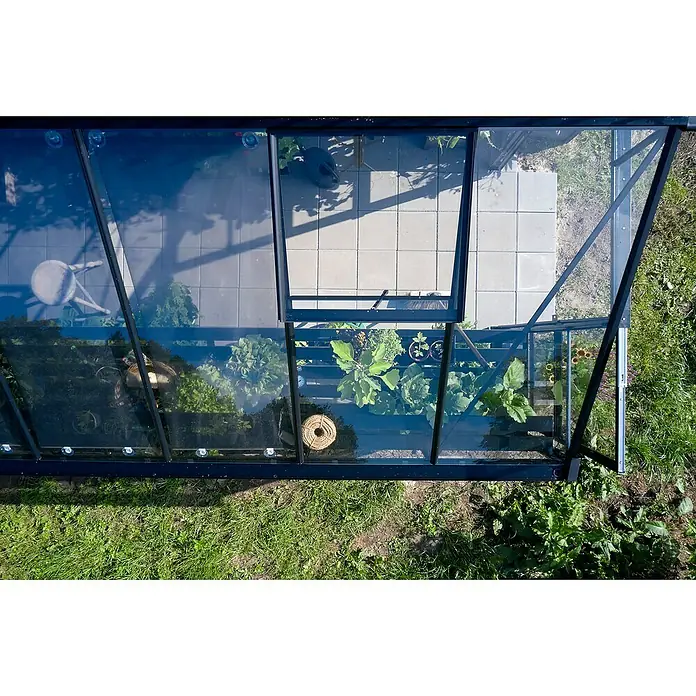 Halls Gewächshaus Blockley 814 - Außenmaß inkl. Dachüberstand (B x T): 256 x 441 cm, Material Seitenwand: Glas, 3 mm, Schwarz - Draufsicht auf ein Gewächshaus mit Pflanzen und Werkzeugen, das für den Gartenbau genutzt wird. Helle Steinplatten bedecken den Boden.