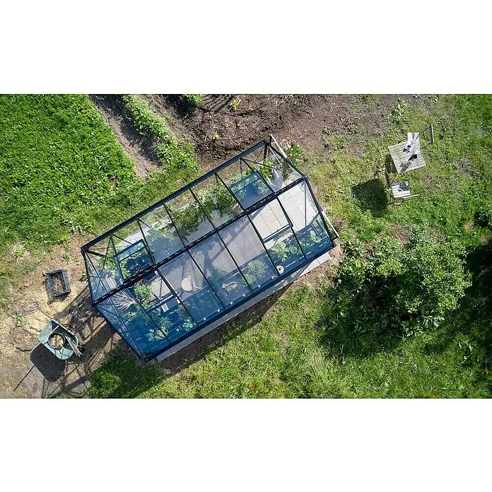Halls Gewächshaus Blockley 814 - Außenmaß inkl. Dachüberstand (B x T): 256 x 441 cm, Material Seitenwand: Glas, 3 mm, Schwarz - Luftaufnahme eines Gewächshauses mit Pflanzen darin, umgeben von einem Garten mit Tisch und Stühlen, sowie Gartengeräten.