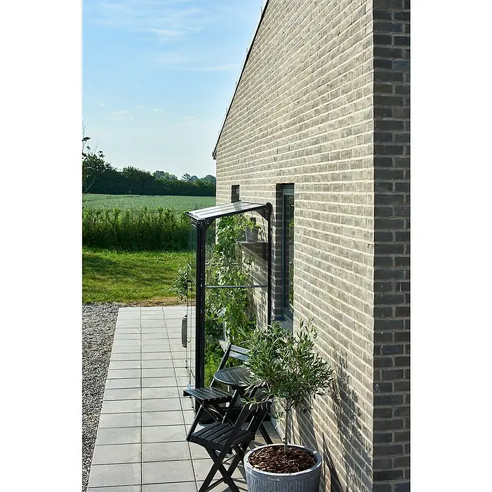 Halls Gewächshaus Qube Lean-to 26 - Außenmaß inkl. Dachüberstand (B x T): 189 x 65 cm, Material Seitenwand: Glas, 3 mm, Schwarz - Das Bild zeigt ein kleines, ansprechendes Gewächshaus mit klaren Glaswänden, das an einer Hauswand steht, ergänzt durch eine stilvolle Sitzgruppe.
