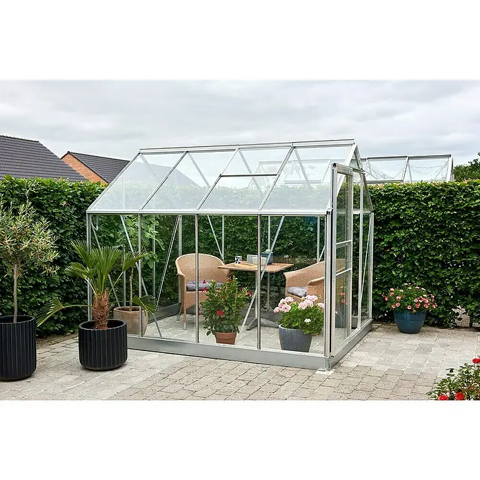 Halls Gewächshaus Popular 86 - Außenmaß inkl. Dachüberstand (B x T): 193 x 257 cm, Material Seitenwand: Glas, 3 mm, Silber - Das Bild zeigt ein Gewächshaus mit Glaswänden und -dach, eingerichtet mit Stühlen und einem Tisch, umgeben von Pflanzen und Büschen.