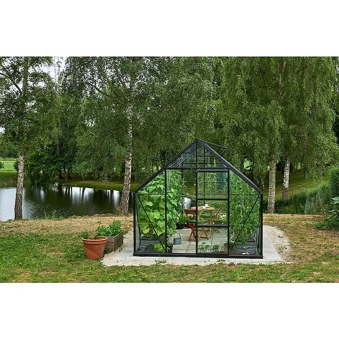 Halls Gewächshaus Universal 128 - Außenmaß inkl. Dachüberstand (B x T): 257 x 384 cm, Material Seitenwand: Glas, Schwarz, Inkl. Fundamentrahmen - Ein Gewächshaus mit Pflanzen steht im Garten, umgeben von Bäumen und einem kleinen Teich. Das Gewächshaus bietet idealen Schutz für die Pflanzen.