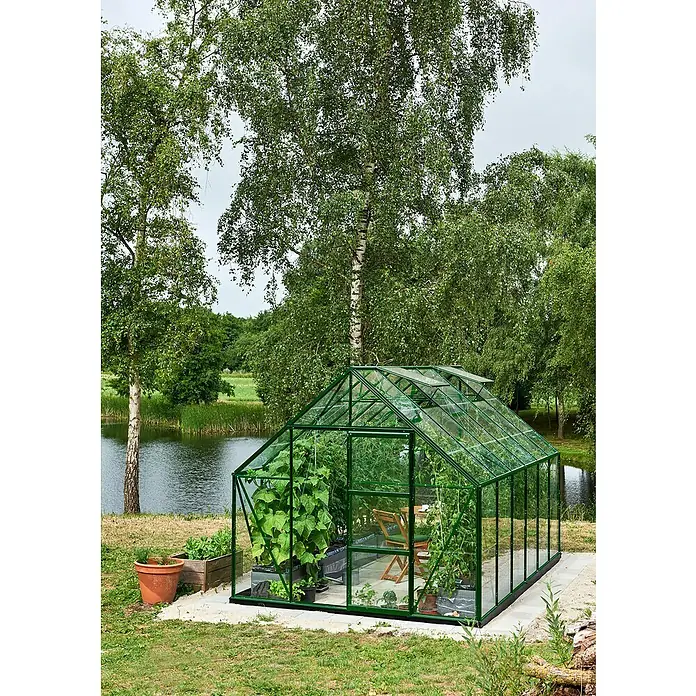 Halls Gewächshaus Universal 128Außenmaß inkl. Dachüberstand (B x T): 257 x 384 cm, Einscheibensicherheitsglas, 3 mm, Grün - Ein grünes Gewächshaus voller Pflanzen steht am Rande eines Gartens, umgeben von Bäumen und einem kleinen Teich.