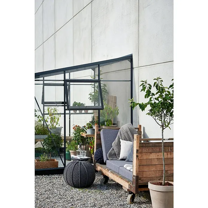 Juliana Gewächshaus Veranda - Außenmaß inkl. Dachüberstand (B x T): 221 x 296 cm, Material Seitenwand: Glas, 3 mm, Anthrazit - Einladende Gartenecke mit einem DIY-Sofa aus Paletten, einem Strickpouf und einem Gewächshaus voller Pflanzen vor einer hellen Wand.