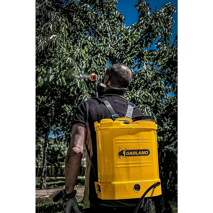 Garland Sulfatadora FUM 312ML-V21 - Presión de funcionamiento: 5 bar - Un hombre con un pulverizador amarillo GARLAND en su espalda rocía un árbol. Es un día soleado y el cielo es azul.