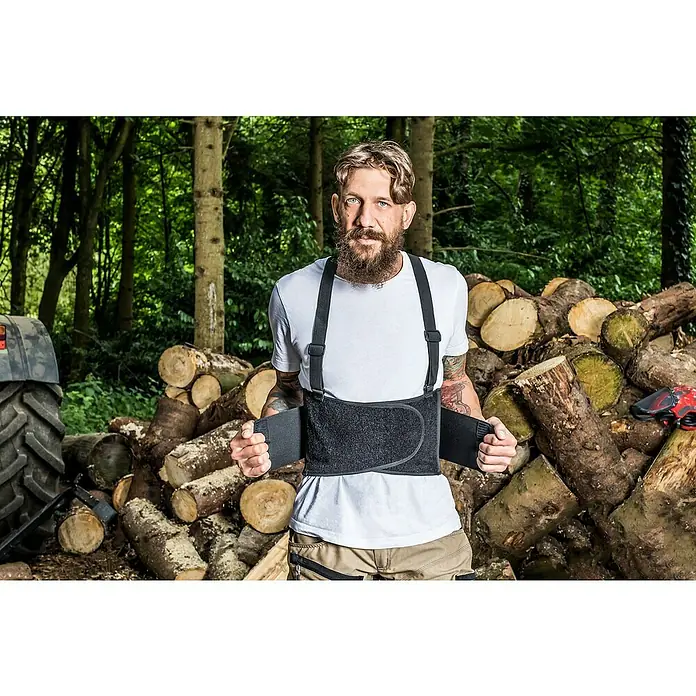 Kneetek Rückenstützgurt BR 108 - XXL, Umfang: 110 m - 138 m - Ein Mann mit Bart trägt einen schwarzen Arbeitsgürtel mit Hosenträgern vor einem Holzhaufen.