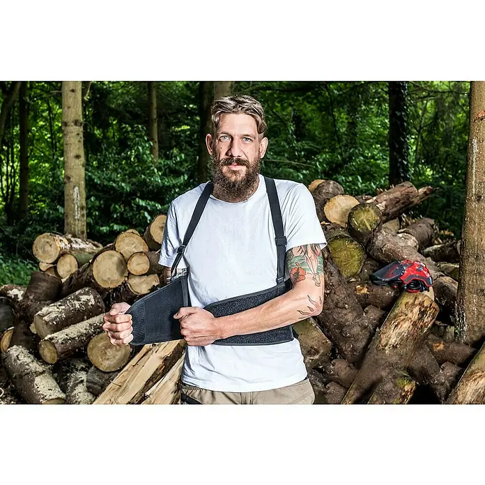 Kneetek Rückenstützgurt BR 108 - XXL, Umfang: 110 m - 138 m - Ein Mann mit Bart und Tätowierung trägt eine Rückenstütze und steht vor einem Holzhaufen im Wald. Die Rückenstütze bietet Halt und Stabilität.