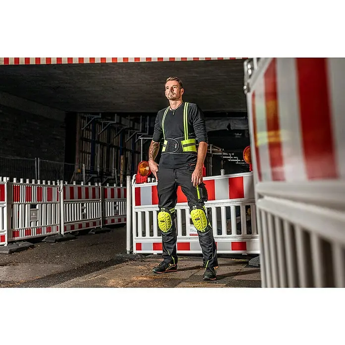 Kneetek Rückenstützgurt HiVis BR 110 - XXL - Ein Bauarbeiter trägt Sicherheitskleidung, darunter Kniepolster und einen Stützgürtel, auf einer Baustelle.