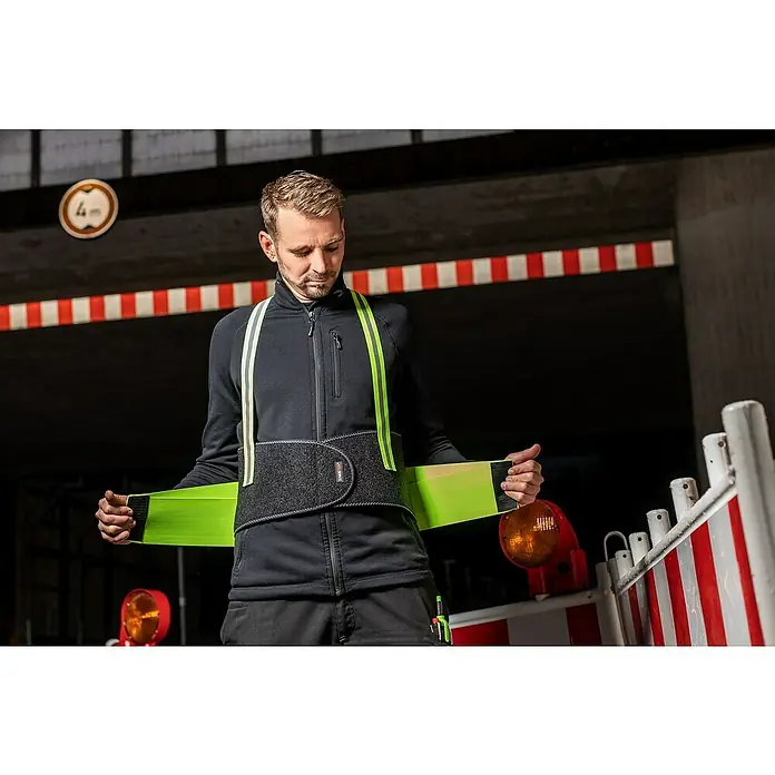 Kneetek Rückenstützgurt HiVis BR 110XL - Ein Mann mit blonden Haaren befestigt eine stützende Arbeitsweste mit neonfarbenen Akzenten vor einem dunklen Hintergrund.