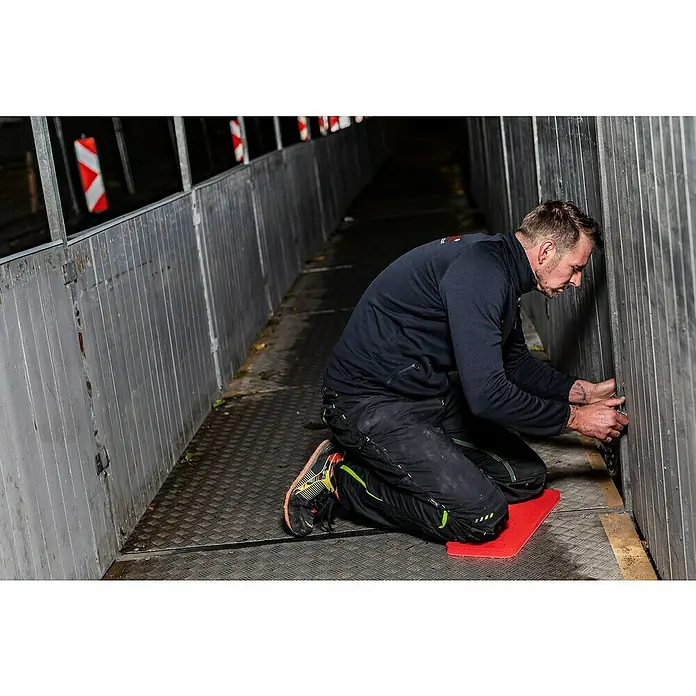 Kneetek Kniekissen RedPad Slim - L x B x H: 450 x 195 x 23 mm - Mann kniet auf einer roten Matte und arbeitet an einer grauen Metallwand. Er trägt eine dunkle Jacke und dunkle Hosen mit neonfarbenen Akzenten.