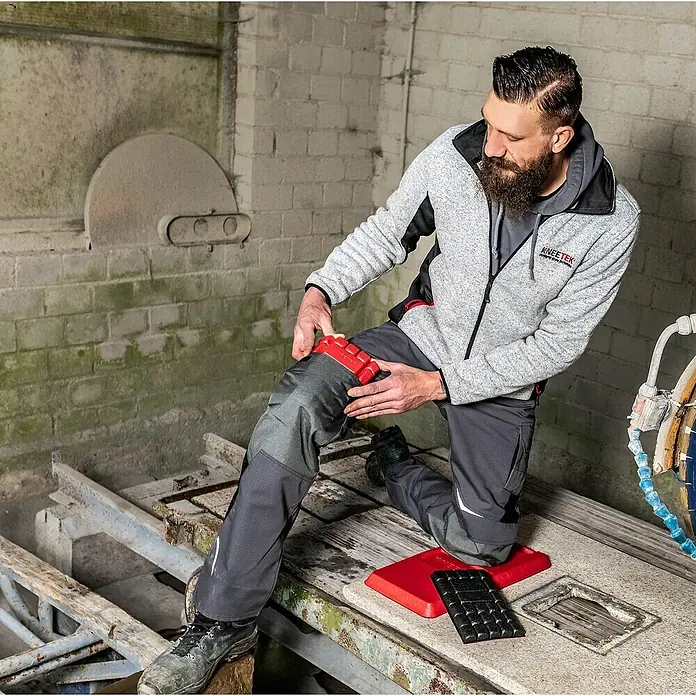 Kneetek Knieschoner MultiPad - Rot, Rechteckig - Ein Mann kniet mit Knieschützern von KNEE-TEK auf einer Werkbank. Die modularen Knieschoner sind rot und schwarz und bieten Komfort und Schutz.