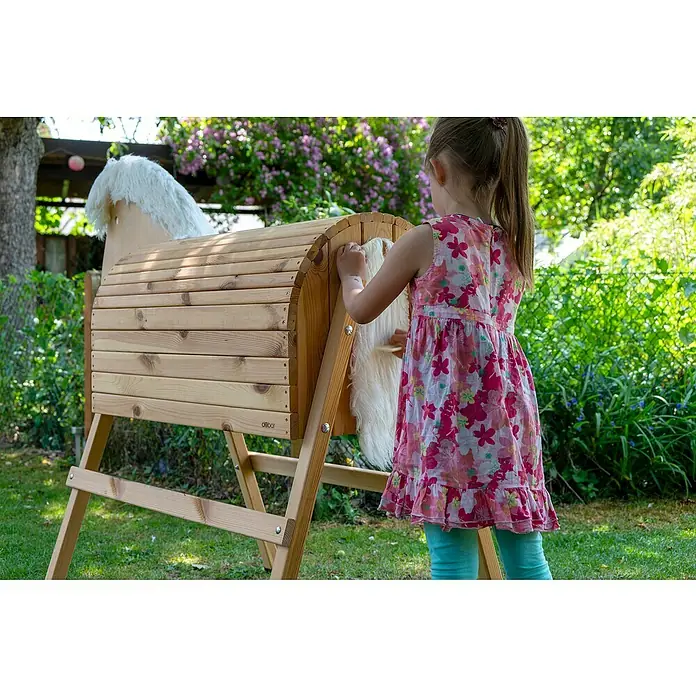 Dobar Holzpferd Bravido - L x B x H: 139 x 78 x 146 cm, Altersempfehlung: 3 Jahre - Ein Kind spielt mit einem hölzernen Spielpferd im Garten. Das Spielpferd ist aus hellem Holz gefertigt und hat eine weiße Mähne.