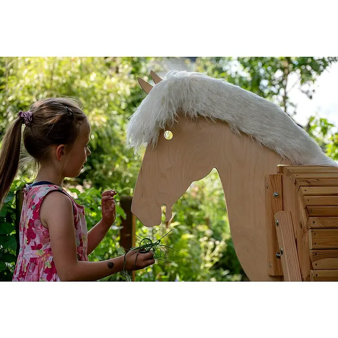 Dobar Holzpferd Bravido - L x B x H: 139 x 78 x 146 cm, Altersempfehlung: 3 Jahre - Ein junges Mädchen füttert ein Holzpferd mit weißer Mähne im Garten, eine idyllische Szene im Grünen.