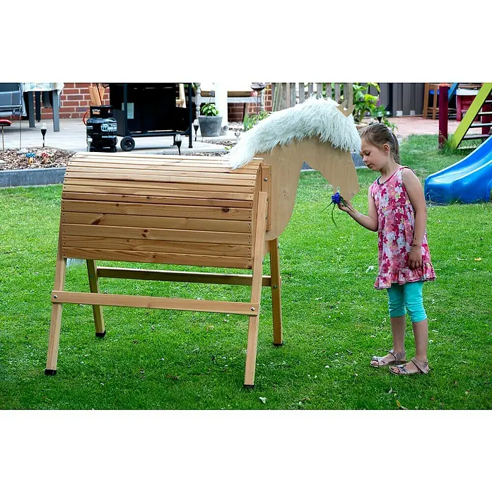 Dobar Holzpferd Bravido - L x B x H: 139 x 78 x 146 cm, Altersempfehlung: 3 Jahre - Mädchen füttert ein hölzernes Spielzeugpferd mit einer blauen Blume im Garten, idyllische Szene mit dem Holzpferd im Mittelpunkt.