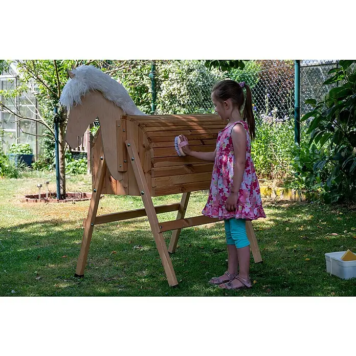 Dobar Holzpferd Bravido - L x B x H: 139 x 78 x 146 cm, Altersempfehlung: 3 Jahre - Ein Mädchen bürstet ein Holzpferd im Garten. Das Holzpferd hat eine weiße Mähne und ist auf einem Holzgestell montiert.