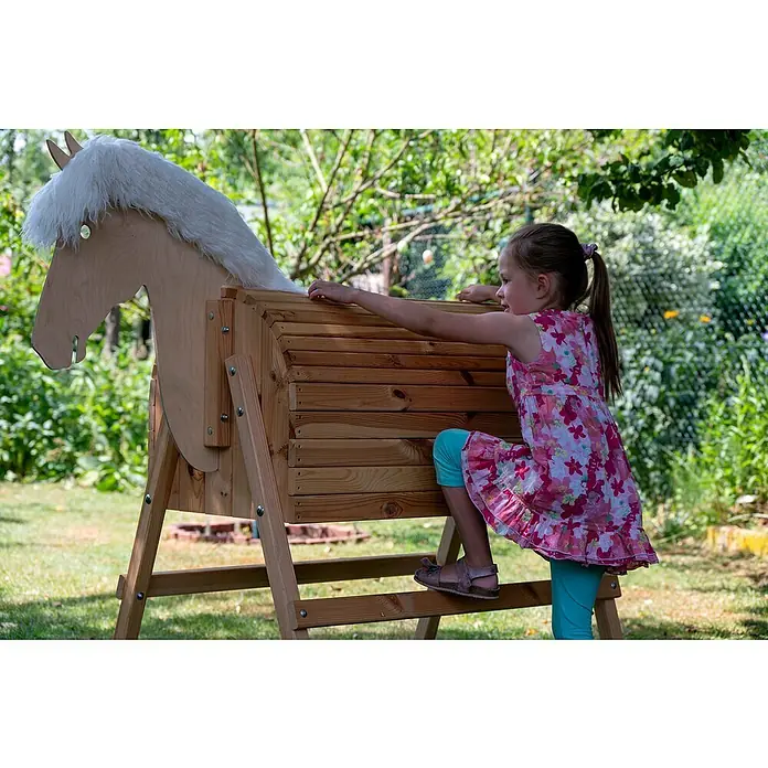 Dobar Holzpferd Bravido - L x B x H: 139 x 78 x 146 cm, Altersempfehlung: 3 Jahre - Ein Mädchen klettert auf ein hölzernes Spielpferd mit weißer Mähne im Garten.