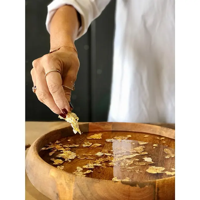 Fleur Resina epoxi Super Transparente - 3,8 l, Transparente - Una mano coloca pan de oro en un cuenco de madera lleno de agua, creando una imagen visual lujosa y brillante.