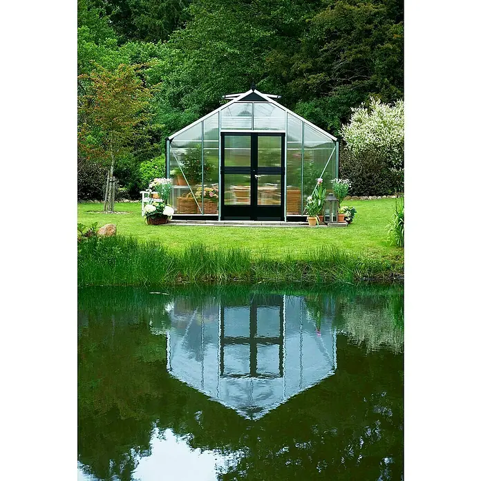 Juliana Gewächshaus Gärtner - Außenmaß inkl. Dachüberstand (B x T): 368 x 583 cm, Material Seitenwand: Kunststoff, 10 mm, Silber/Schwarz - Ein transparentes Gewächshaus mit dunklen Türen steht am Ufer eines Sees und spiegelt sich malerisch im Wasser. Umgeben von üppigem Grün.