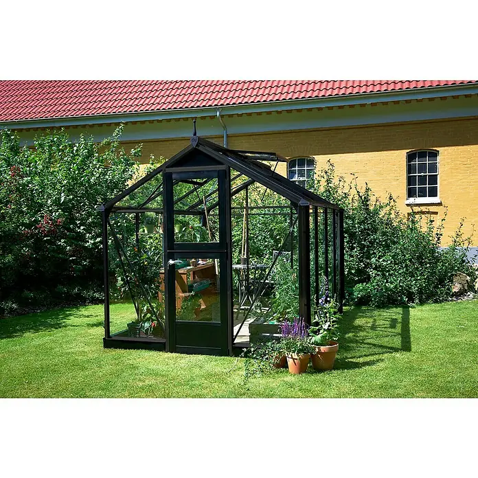 Juliana Gewächshaus Compact - Außenmaß inkl. Dachüberstand (B x T): 224 x 295 cm, Material Seitenwand: Glas, 3 mm, Anthrazit/Schwarz - Ein elegantes schwarzes Gewächshaus mit Glasscheiben steht inmitten eines grünen Gartens, umgeben von Pflanzen und Blumen.