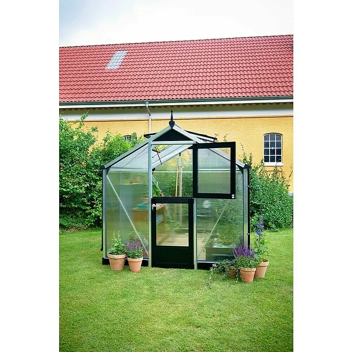 Juliana Gewächshaus Compact - Außenmaß inkl. Dachüberstand (B x T): 224 x 295 cm, Material Seitenwand: Kunststoff, 10 mm, Silber/Schwarz - Gewächshaus mit geöffneter Tür und Fenster umgeben von Blumentöpfen, vor einem gelben Gebäude mit rotem Dach.