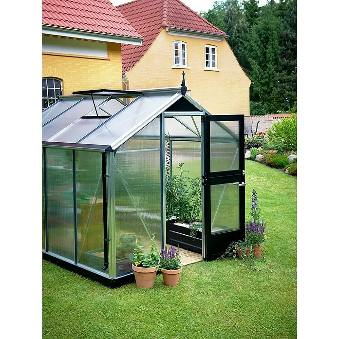 Juliana Gewächshaus Compact - Außenmaß inkl. Dachüberstand (B x T): 224 x 295 cm, Material Seitenwand: Kunststoff, 10 mm, Silber/Schwarz - Nahaufnahme eines Gewächshauses mit geöffneter Tür, umgeben von üppigem Grün und Blumen. Das Gewächshaus bietet eine ideale Umgebung für Pflanzen.