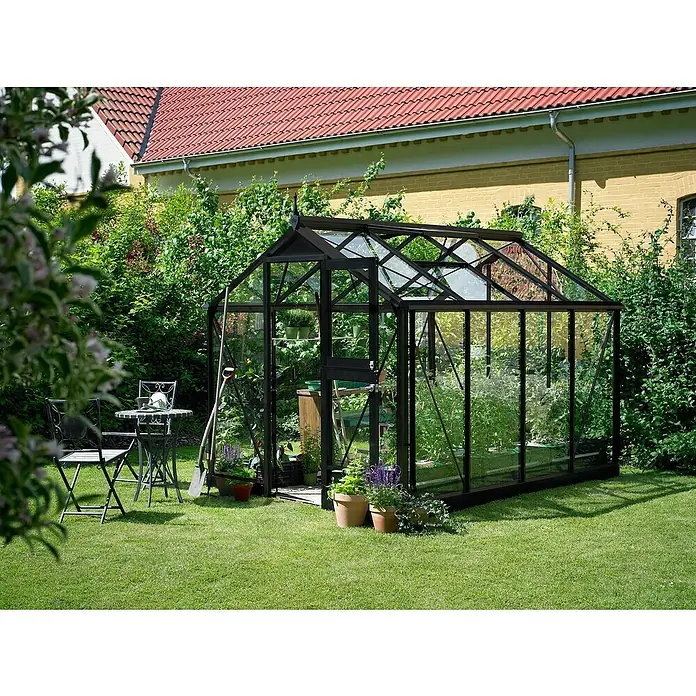 Juliana Gewächshaus Compact - Außenmaß inkl. Dachüberstand (B x T): 224 x 295 cm, Material Seitenwand: Glas, 3 mm, Anthrazit/Schwarz - Ein elegantes Gewächshaus aus Glas und Metall steht im Garten, umgeben von Grünpflanzen und Blumen.